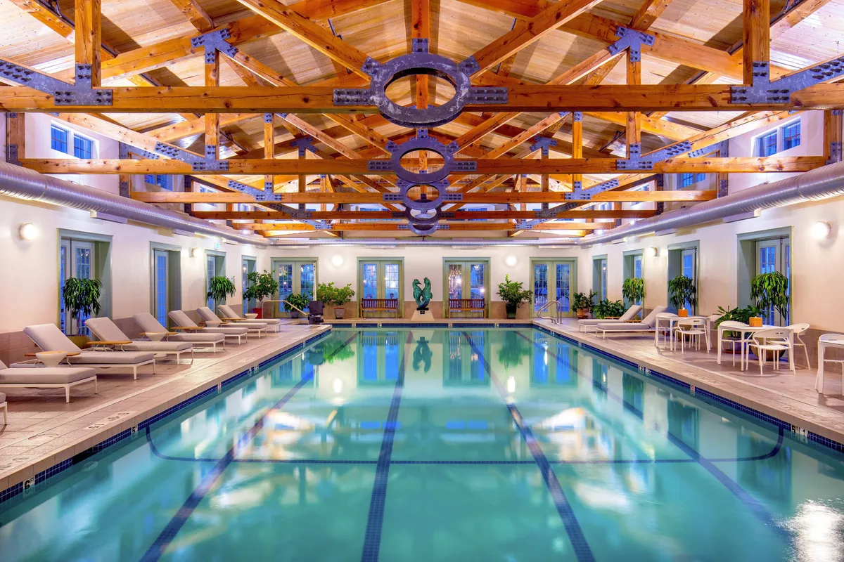 A serene indoor swimming pool area framed by elegant wooden beams, lush greenery, modern seating, and a striking sculpture. The tranquil ambiance suggests opportunities for relaxation, socializing, and wellness activities such as yoga or meditative reflection.
