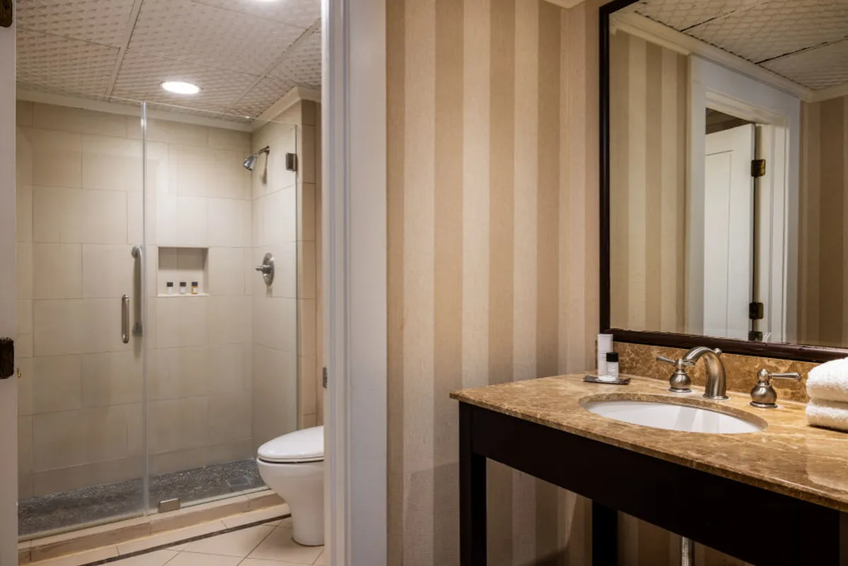 A well-appointed bathroom featuring a glass shower enclosure, marble countertop, and striped wallpaper. The vanity mirror and overall layout suggest a comfortable, ADA-friendly environment suitable for all guests. The space exudes a sense of pampering and modern comfort.
