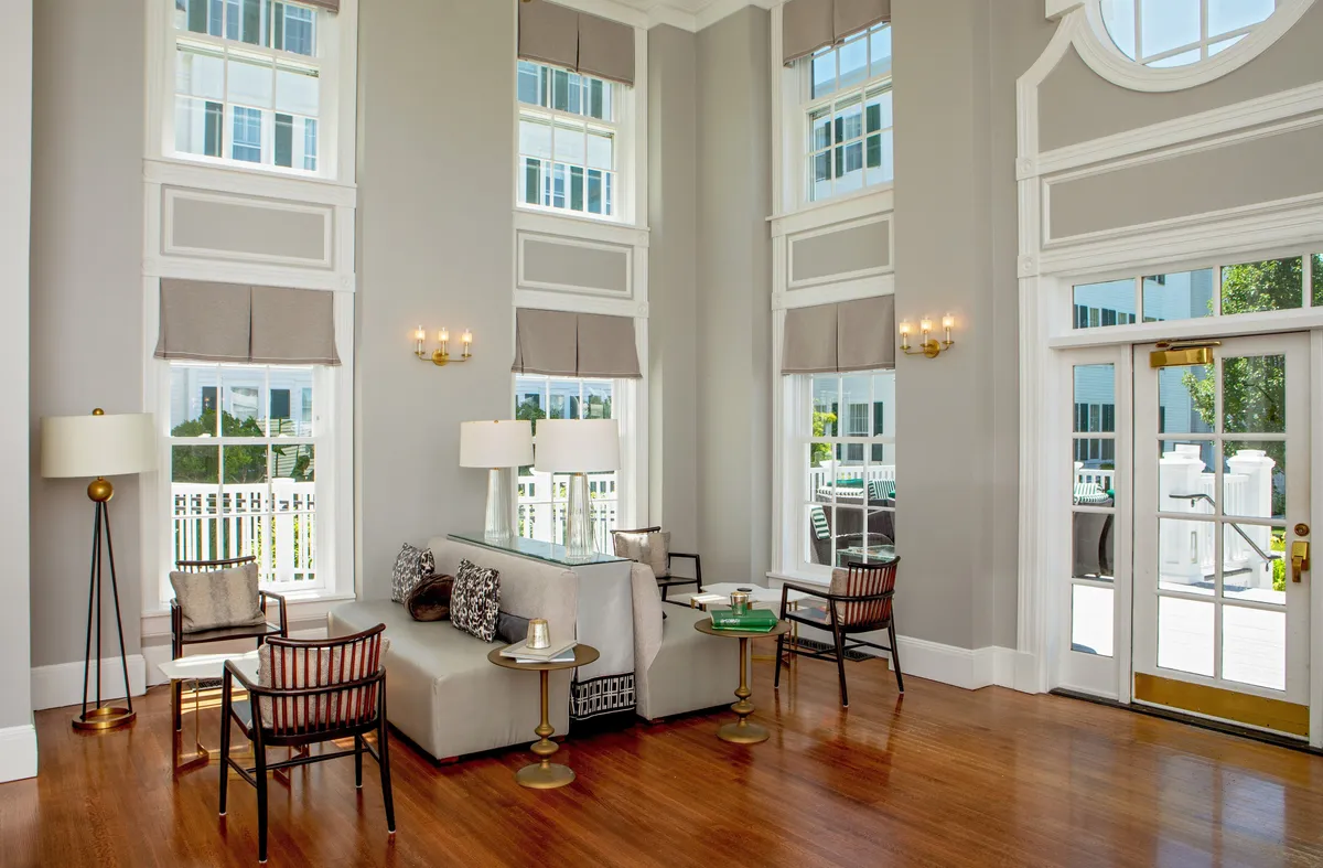 A warmly lit guest room featuring a comfortable seating area with a decorative lamp and coffee table, surrounded by large windows that frame the outdoors. The space is individually decorated, with plush furnishings inviting relaxation and ample natural light creating a tranquil atmosphere.