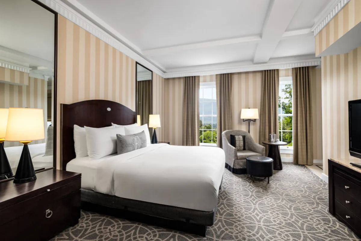 The image showcases a serene, light-filled studio interior at The Equinox Golf Resort & Spa. A plush king-sized bed sits invitingly beside floor-to-ceiling windows that bathe the room in natural light. A stylish armchair offers a cozy spot for reading or relaxing, and elegant decor creates a refined yet comfortable atmosphere. The mirrored wall adds to the sense of spaciousness, making the studio feel open and airy.