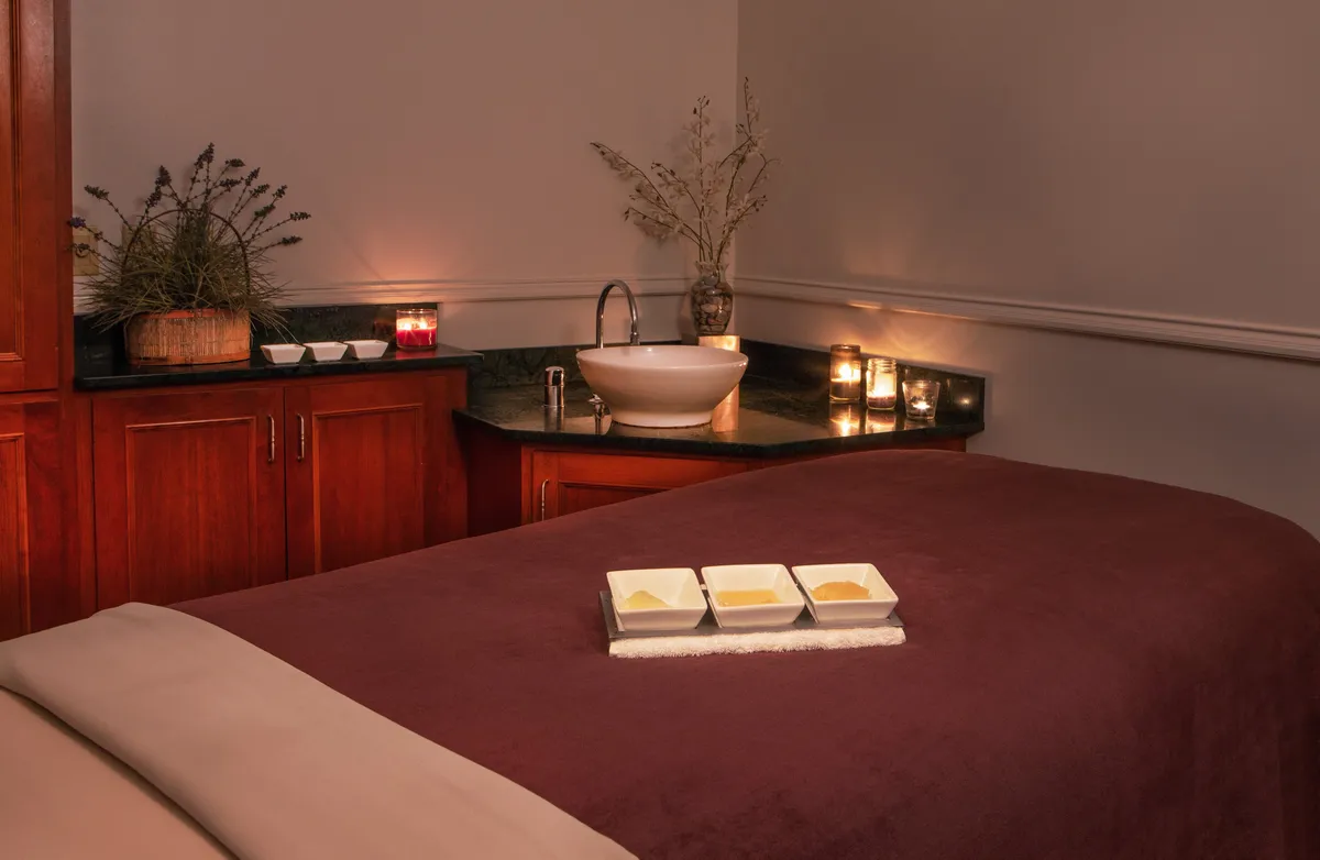 A tranquil spa treatment room with a plush bed, decorative bowls filled with creams, and ambient candlelight, offering a serene environment designed for relaxation and wellness therapies.
