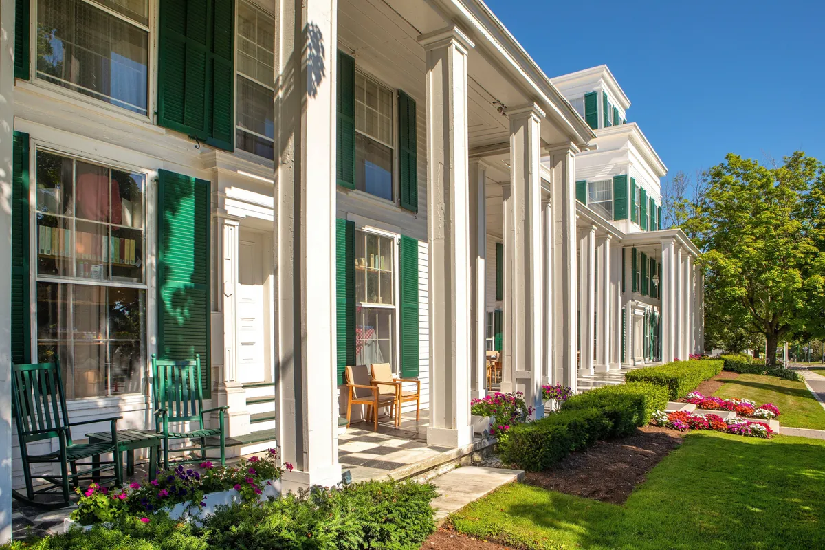This exterior image presents The Equinox Golf Resort & Spa's inviting front porch, adorned with classic green shutters, sturdy white columns, and carefully tended flower beds. Rocking chairs line the porch, offering a tranquil spot to unwind and enjoy the serenity of the gardens and outdoor surroundings, with hints of mountain vistas beyond.