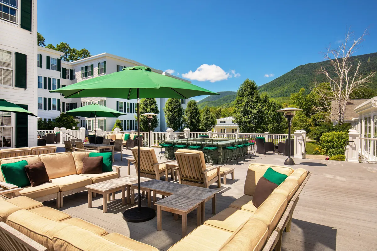 An inviting outdoor dining scene unfolds beneath green umbrellas, with plush sofa seating arranged to take full advantage of breathtaking mountain views. The terrace is set for relaxed socializing, and the mood is tranquil, perfect for outdoor brunches, sunset drinks, or evening gatherings warmed by heaters.