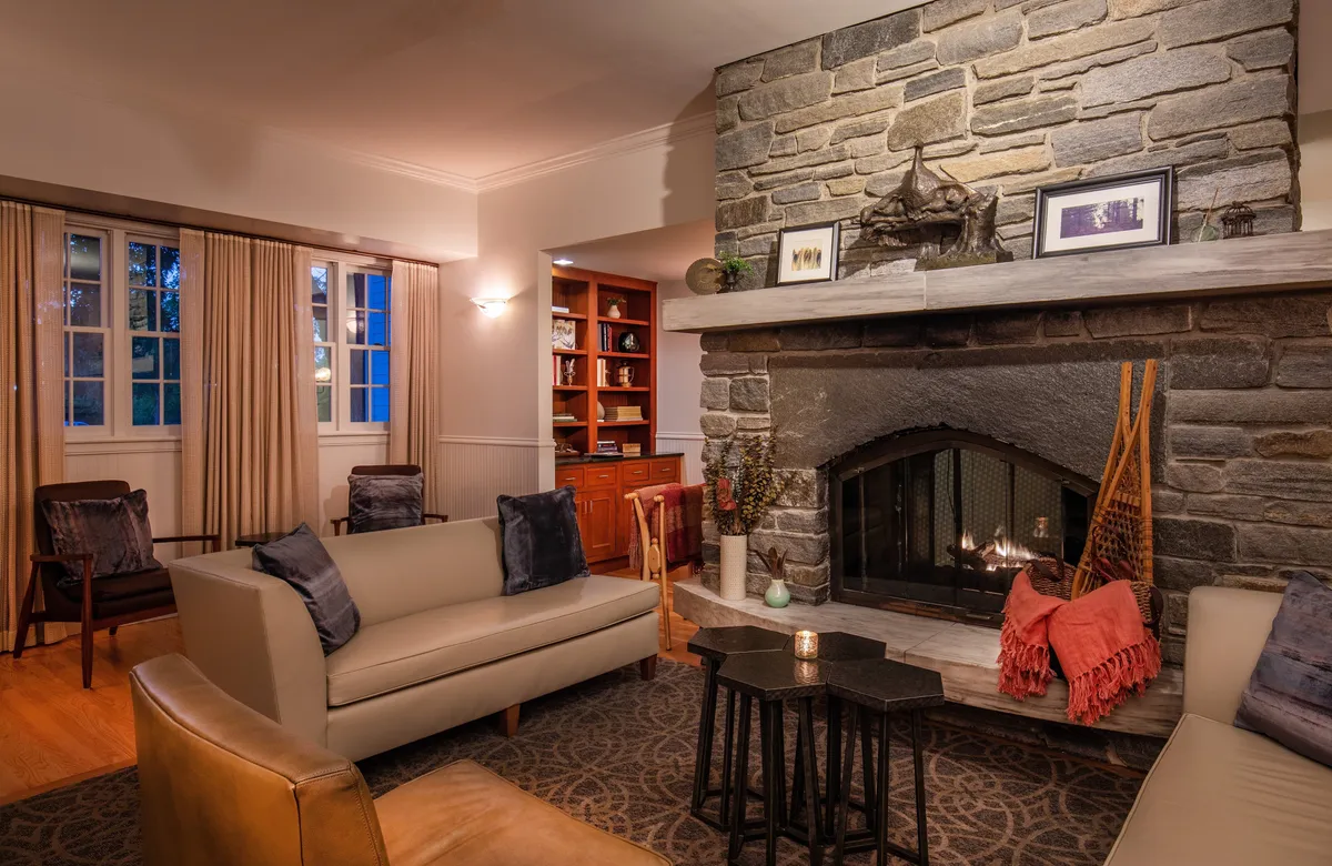A cozy interior lounge featuring a crackling fireplace, plush sofas, a bookshelf lined with books, and decorative items that evoke comfort and relaxation. The scene is softly lit, inviting guests to unwind and socialize in a refined yet homely atmosphere.