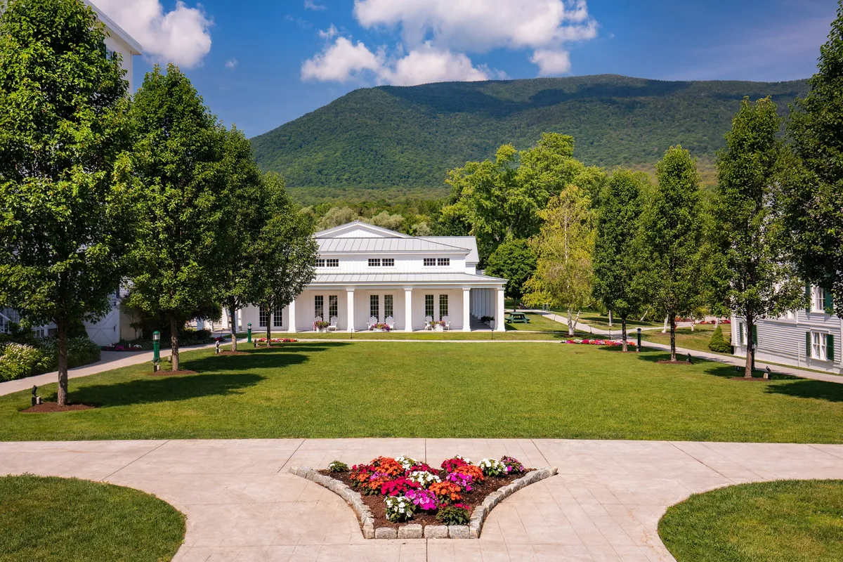The impressive main resort building set amidst colorful flower beds and mature trees, with mountains rising dramatically in the background. The grounds are calm and picturesque, hinting at outdoor leisure and gatherings.