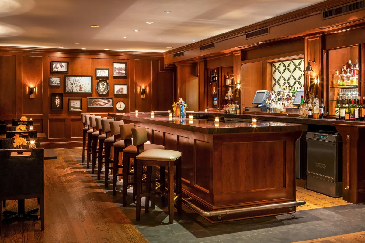 This image features a cozy interior bar area, complete with a polished bar counter, comfortable stools, and walls adorned with artwork. Liquor bottles line the shelves, hinting at an extensive drink selection. The atmosphere is lively, with people socializing, enjoying drinks, and relaxing in a warm, inviting space. Signature cocktails and tasting events are highlighted, contributing to an engaging social scene.